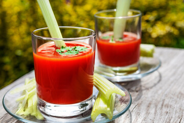 Two glasses of tomato juice with parsley and celery decorations, stand in the open air on wooden boards, the rays of the sun shine, shallow depth of field, selective focus. The concept of natural