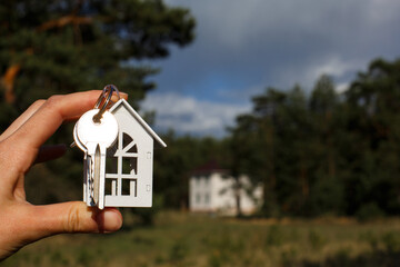 Wooden key chain and keys in hand on the background of an unfinished house. Dream of moving, the cottage is in a rural location and completion of the project. Space for text