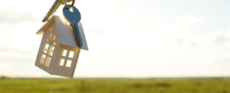 White wooden figure of a house and keys in hand against the background of the sky and field. Dream of your home, building a cottage in the countryside, plan and design, delivery of the project, moving