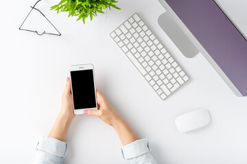 Woman’s hands using smart phone with empty screen on white table. Office desktop. Flat lay