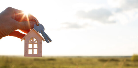 White wooden figure of a house and keys in hand against the background of the sky and field. Dream of your home, building a cottage in the countryside, plan and design, delivery of the project, moving