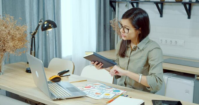 Young woman teacher teaching English or alphabet online using a laptop while looking at her watch and making notes in a notebook setting a date for a new lesson at home. Online education.