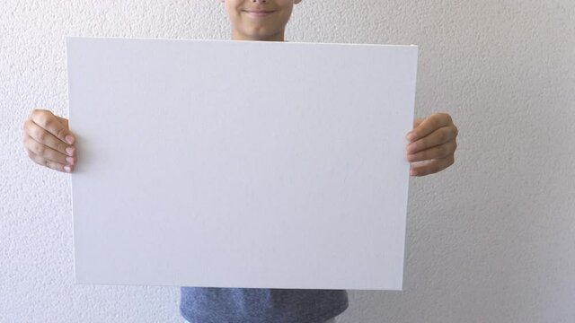 Boy holding in his hands and show blank empty white mock up board poster. Grey wall on background. Close up