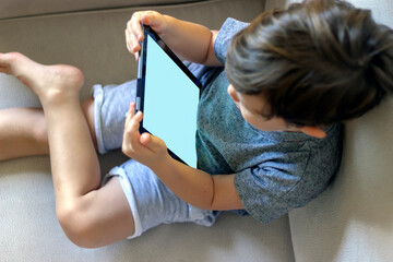 High angle shot of smiling boy watching film with help of his tablet while relaxing on sofa.Smarthone in kids hands.Toddler playing with tablet at home.Boy looking on digital tablet.Technology concept