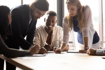 Diverse associates gathered in boardroom leaned over desk writing project plan and ideas do...