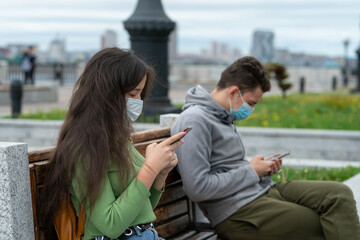social distance, a guy and a girl are sitting on a bench at a distance from each other with masks on their faces and with phones in their hands