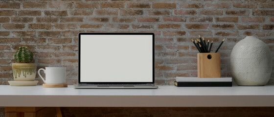 Contemporary worktable with mock-up laptop, stationery, cup and decorations on white desk with brick wall
