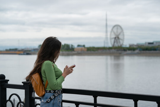 a girl stands on the embankment of the river, looks at the phone.  on the other side of the river the Chinese city of Heihe, a Ferris wheel.