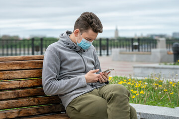 Obraz premium Young man on the street sits on a bench in a medical mask and with a phone in his hands.