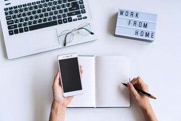 Flat lay of woman hand writing on notebook while using smartphone on white desk with computer laptop, glasses and work from home word on light box. Copy space. Quarantine, new normal  concept.