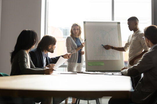 Diverse business people listen african ethnicity mentor caucasian woman trainer make presentation on flip chart, serious investor during negotiations group meeting ask question regarding data on graph