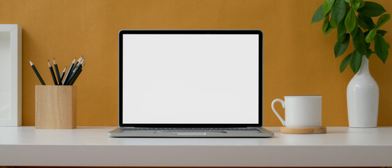 Creative worktable with mock-up laptop, stationery, cup and decorations on white table with yellow wall