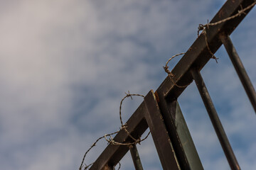 Closed rusty iron gate with barbed wire on top and blue sky with clouds on background