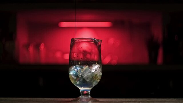 Pouring fresh alcoholic drink into the glass with ice cubes on the bar counter. Stock footage. Close up of process of cocktail preparation, adding blue syrup into the glass.