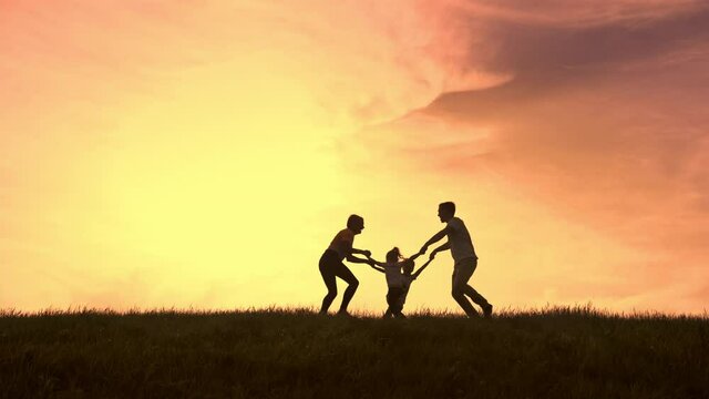 In general, the silhouettes of parents with two children in the background of sunset hold hands and swirl gaily in a circle, then they hug and raise the children in their arms.
