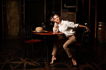 Beautiful theatrical actress woman with retro fashion style on the stage siting near wooden table with glass of red wine and vintage hat. 