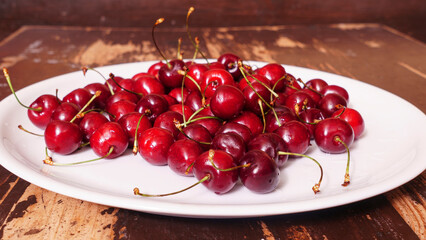 A lot of fresh sweet cherry fruit berries with water drops. Close-up on the plate on wooden background. Close-up of pile of ripe cherries. Large collection of fresh red cherries. Ripe cherries texture