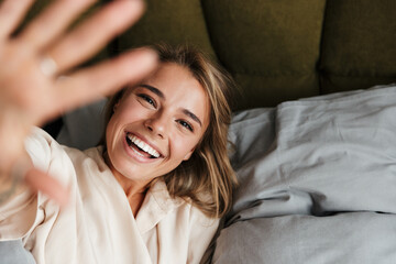 Image of laughing woman making fun and waving hand while lying in bed