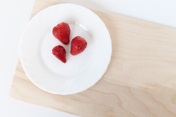 three red strawberries on a white plate on the brown wooden cutting board on the white background