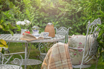 Cozy tea party at garden with book, teapot, candle, flowers, blanket. Beautiful metal lacy chairs and table. cosiness. tranquility. relaxation. Sunlight. Nobody