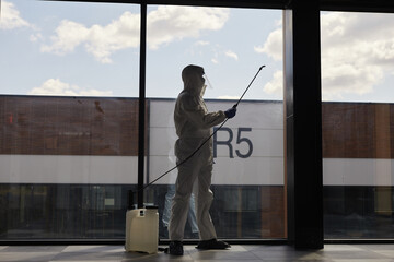 Backlit full length portrait of worker wearing protective suit spraying chemicals during disinfection or cleaning, copy space