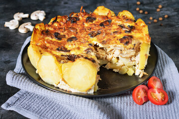 Homemade Lorraine pie with potatoes, chicken, mushrooms, cheese, onions and seasonings. In a black plate, on a dark table with  tomatoes, a gray napkin, champignons and black pepper and bay leaf.