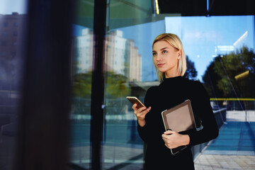 Portrait of beautiful young female holding mobile phone in hands while strolling urban park.