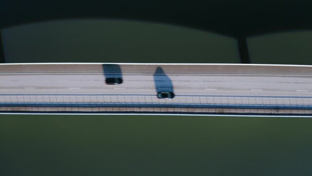 Aerial top down view and flying up above highway bridge over river; two lanes, one direction, very low car traffic, long shadows