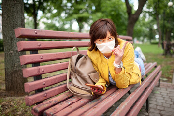 Funny youthful woman lies on her stomach in park bench with a protective medical mask on sunny warm summer day. Concept of humor during the coronovirus pandemic