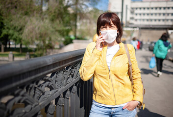 Fototapeta premium Funny middle-aged woman in a white protective mask talking on a smartphone while walking around the city on a warm spring day. Coronovirus pandemic and remote communication concept