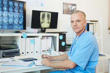 Portrait of handsome mature doctor looking at camera while posing at workplace in modern clinic interior with CT scans in background, copy space
