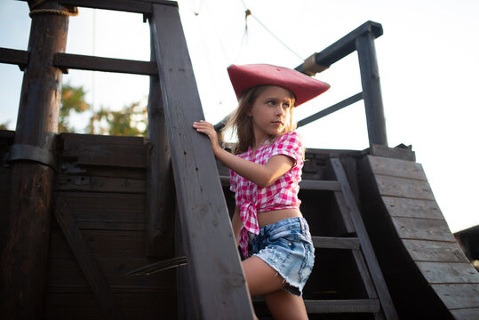 Serious Funny Little Girl Pirate In A Plaid Shirt And Denim Shorts Holds On To The Cords Of The Ship. Concept Kids In Adventure Seekers