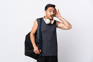 Young sport man with sport bag isolated on white background listening to something by putting hand on the ear