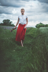 Beautiful young woman girl in red skirt holding herbs bouquet and walking on grass between fields in dark moody time