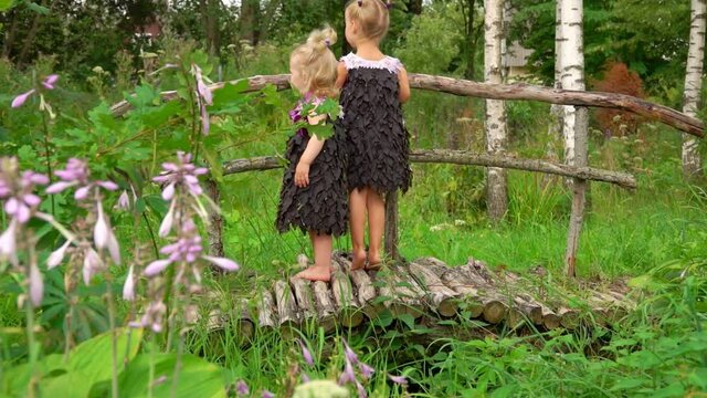 Two little girls in a purple fantasy dresses made of leaves and flowers are standing on a wooden bridge in the forest