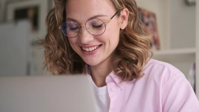 A happy pleased young blond woman wearing glasses is using her laptop computer sitting at the table indoors