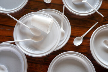 Platos, vasos y cubiertos de p&aacute;stico blanco desechables esparcidos en una mesa de madera oscura.
