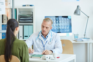 Portrait of mature male doctor writing on clipboard while sitting at desk and consulting female patient in modern clinic, copy space