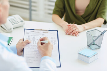 Close up of unrecognizable doctor writing on clipboard and filling patients report while consulting young woman in modern clinic, copy space