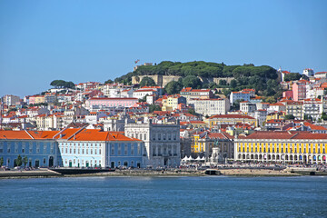 Fototapeta na wymiar Multi coloured buildings of the old city of Lisbon. São Jorge Castle looking down on the city Praça do Comércio town square in the foreground. View from across the Tagus Estuary, Portugal.