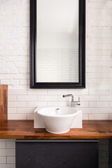 Big mirror with dark frame hanged on white painted and tiled brick wall over wooden counter around ceramic sink in light washroom with interior design in loft style