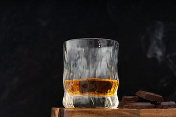 Glass of golden whiskey and dark chocolate pieces on wooden table against dark background