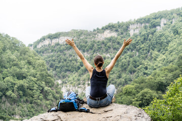 A sports woman sits on a peak of a mountain with a backpack and throws up her hands to freedom. Digital detox and mental health concept. Positive emotions and satisfaction with a healthy lifestyle