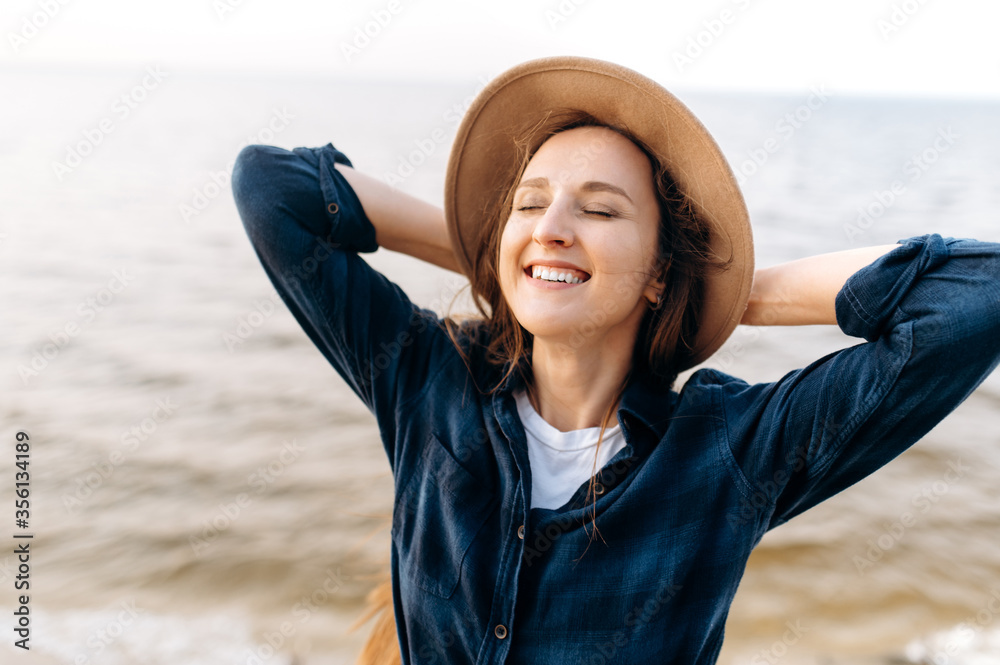 Wall mural Close-up portrait of a gorgeous smiling caucasian young woman whose posing in a hat near the sea in a positive mood