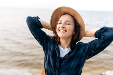 Close-up portrait of a gorgeous smiling caucasian young woman whose posing in a hat near the sea in a positive mood