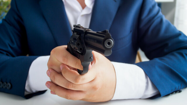 Closeup image of businessman in suit sitting behind desk and aiming with gun