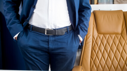 Stylish young businessman in blue suit standing in modern office with brown leather armchair