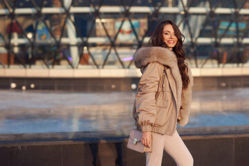 Stylish young woman wearing casual winter clothes and coat with fur hood standing or walking at city street on a sunny day. Fashionable girl