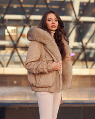 Stylish young woman wearing casual winter clothes and coat with fur hood standing or walking at city street on a sunny day. Fashionable girl