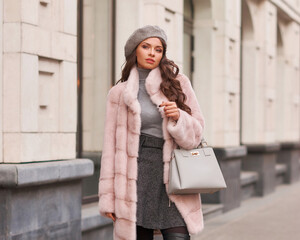 Elegant rich woman in pink fur coat and gray beret walking city street. Outdoor life style portrait of fashionable lady